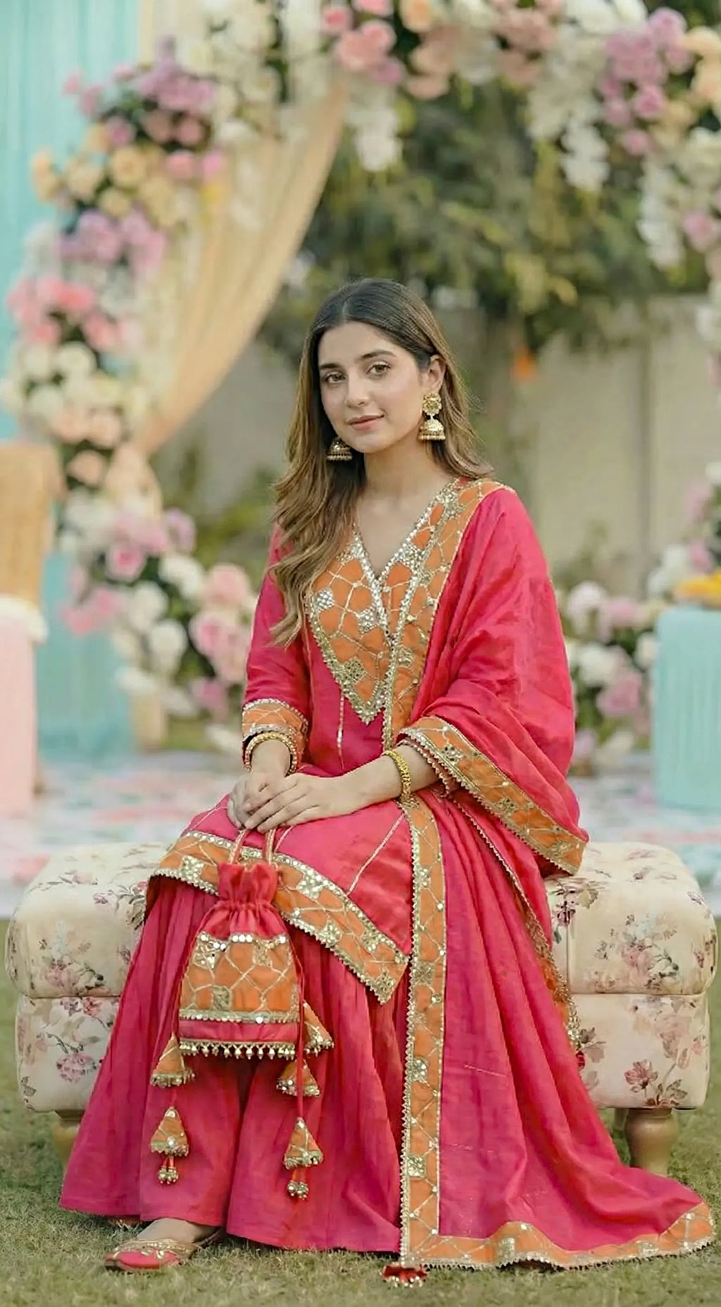 Woman in a hot pink Anarkali Sharara set with gota sequin work and mirror detailing by Karchobi, seated outdoors amid floral decor.