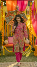 Woman in a pink floral Zari embroidered kurta pant set by Karchobi, holding a colorful umbrella at an outdoor festive event with vibrant drapes and marigold decorations.